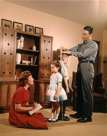 1950s 1960s FAMILY MAN FATHER WOMAN MOTHER BOY SON GIRL DAUGHTER MEASURING AND COMPARING HEIGHT OF SISTER AND BROTHER IN LIVING ROOM Stock Photo - Rights-Managed, Code: 846-02794441