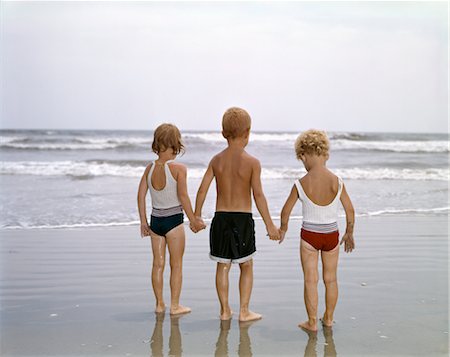 1960s YOUNG CHILDREN BOY AND TWO GIRLS IN BATHING SUITS HOLDING HANDS ON THE BEACH LOOKING AT THE OCEAN WAVES Stock Photo - Rights-Managed, Code: 846-02794472