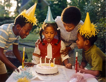 1970s AFRICAN AMERICAN MOTHER 3 CHILDREN LITTLE GIRL BLOWING OUT CANDLES BIRTHDAY CAKE 2 BOYS IN YELLOW PAPER PARTY HATS Stock Photo - Rights-Managed, Code: 846-02794515