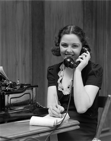 1930s SMILING YOUNG WOMAN RECEPTIONIST SECRETARY SITTING AT DESK IN OFFICE TALKING ON TELEPHONE Stock Photo - Rights-Managed, Code: 846-02795815