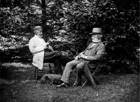 TURN OF THE CENTURY TWO MEN SITTING OUTSIDE IN CHAIRS AMONG TREES SMOKING CIGARS TALKING DOG LAYING AT THEIR FEET Stock Photo - Rights-Managed, Code: 846-02795978