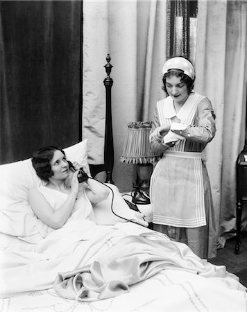 1920s 1930s TWO WOMEN IN BEDROOM MAID LOOKING AT WRIST WATCH OTHER WOMAN IN BED TALKING ON PHONE HAND HELD OVER MOUTHPIECE Stock Photo - Rights-Managed, Code: 846-02797626