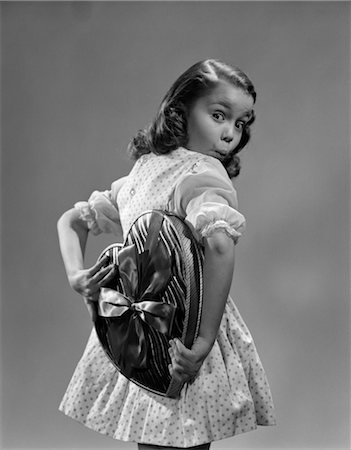 1950s YOUNG GIRL HOLDING HEART SHAPED CANDY BOX BEHIND BACK WITH SURPRISED EXPRESSION WEARING POLKA DOT DRESS Stock Photo - Rights-Managed, Code: 846-02797729
