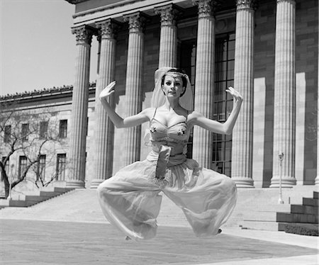 1960s WOMAN IN MIDDLE EASTERN BELLY DANCE COSTUME JUMPING IN FRONT OF BUILDING WITH GREEK STYLE COLUMNS OUTDOOR Stock Photo - Rights-Managed, Code: 846-02792434