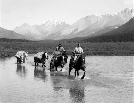 simsearch:846-02793153,k - 1930s TWO WOMEN MAN RIDING HORSEBACK THROUGH SHALLOW WATER TWO EXTRA HORSES CARRY CAMPING EQUIPMENT MOUNTAINS IN BACKGROUND Stock Photo - Rights-Managed, Code: 846-02793219