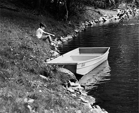 1960s SIDE VIEW OF BOY SITTING ON BANK OF LAKE NEXT TO ROWBOAT IN WATER Stock Photo - Rights-Managed, Code: 846-02793531