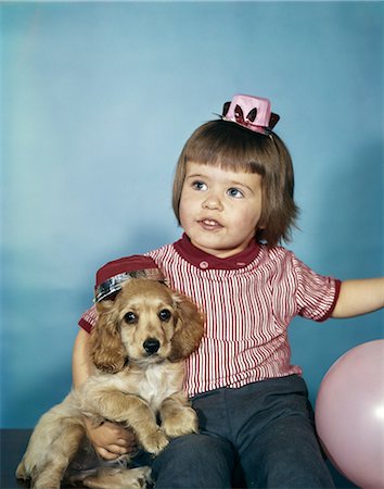 1950s 1960s LITTLE GIRL IN PARTY HAT SITTING HOLDING A COCKER SPANIEL PUPPY WITH BALLOONS STUDIO Stock Photo - Rights-Managed, Code: 846-03164101