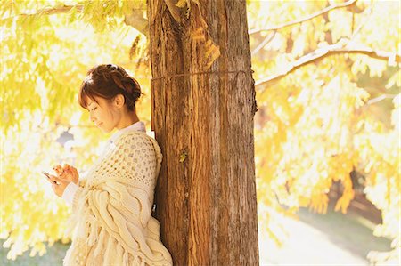 poncho - Japanese woman in a white cardigan with a Smartphone leaning against a tree Stock Photo - Rights-Managed, Code: 859-06404987