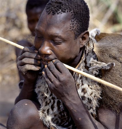 simsearch:862-03821015,k - A Hadza hunter wearing as genet cat skin cape straightens a new arrow shaft in his teeth. The Hadzabe are a thousand-strong community of hunter-gatherers who have lived in the Lake Eyasi basin for centuries. They are one of only four or five societies in the world that still earn a living primarily from wild resources. . Foto de stock - Con derechos protegidos, Código: 862-03355119