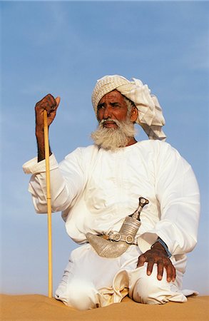 A Bedouin man kneels on top of a sand dune in the desert . He wears the traditional Omani long white cloak or dish dash,a turban,a ceremonial curved dagger (khanjar) and holds a short camel stickOman 7John Warburton-Lee Stock Photo - Rights-Managed, Code: 862-03360155