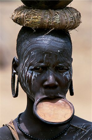 A woman of the Mursi tribe. Once married Mursi women pierce their lower lip and stretch it by inserting  increasingly large plugs until they can wear a clay lip plate. The size of the lip plate reflects the bride price paid by their husband. Within the Omo Valley, the Mursi have a reputation for being extremely fierce and aggressive but also for their skill at making pots. This women carries a pot Stock Photo - Rights-Managed, Code: 862-03820425