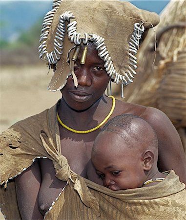 A Mursi mother and child.The mother shades her shaven head from the sun with a small decorated leather apron.The Mursi speak a Nilotic language and have affinities with the Shilluk and Anuak of eastern Sudan. They live in a remote area of southwest Ethiopia along the Omo River. Stock Photo - Rights-Managed, Code: 862-03820473
