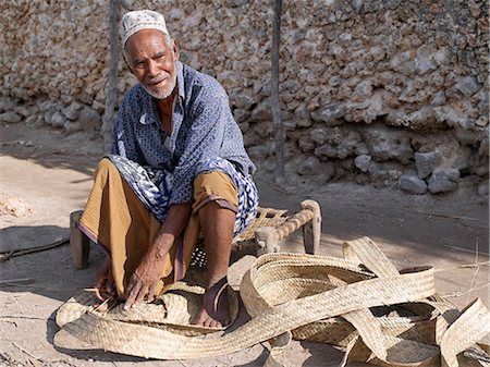 Assis sur un tabouret de côte typique, un homme de Swahili points lanières de feuilles de palmier tissé dans un matelas dans une des ruelles de Pates.Tous les bâtiments en pierre sont construits de corail chiffon avec des toits de makuti, qui sont un type de chaume fabriqué à partir de feuilles de cocotier. Photographie de stock - Rights-Managed, Code: 862-03820756