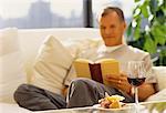Mature homme assis sur un canapé lecture livre avec vin et fromage