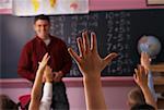 Male Teacher Looking at Children With Hands Raised in Classroom