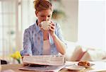 Woman Sitting at Table Reading Newspaper, Drinking from Mug