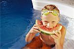Garçon en maillot de bain et lunettes mangeant melon d'eau dans la piscine