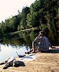 Back View of Man Sitting on Dock Using Cell Phone with Laptop and Dog, Bala, Ontario, Canada