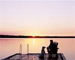 Rückansicht des Mann sitzt im Stuhl auf Dock mit Hund bei Sonnenuntergang Bala, Ontario, Kanada