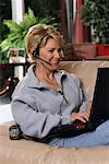 Mature Woman Sitting on Sofa Using Telephone Headset and Laptop Computer