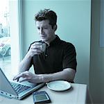 Man Sitting at Table by Window in Cafe, Using Computer, Holding Cup