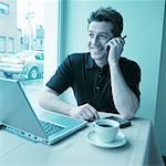 Man Using Cell Phone in Cafe with Laptop Computer on Table