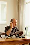 Man Sitting at Desk Holding Paper and Using Cell Phone