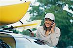 Woman Talking on Cellular Phone, Tying Kayak to Car Roof