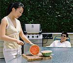 Woman Slicing Watermelon and Man Relaxing