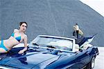 Frauen im Bikini auf der Motorhaube des Muscle-Car mit Mann, Surfboard im Hintergrund hält
