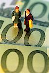 Toy businessman and businesswoman standing on top of Euro one hundred bank notes, close-up