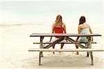 Rear view of a girl and a teenage girl sitting on a picnic table on the beach