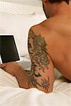 Man lying on bed, using laptop (close-up on upper arm with tattoo)