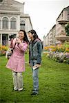 Young Asian woman with a camera standing next to a younger woman in front of a park and is laughing, selective focus