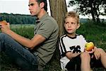 Father and son leaning against a tree eating apples
