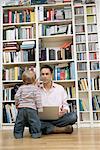 Father with a laptop and a mobile phone sitting in front of a shelf son standing in front of him