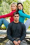 Portrait of a young man sitting on a bench with two young women sitting back to back behind him