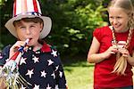 Portrait of Boy Wearing Stars and Stripes Top and Hat, Blowing Noisemaker Horn, Girl Watching