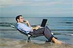 Side profile of a young man sitting on a lounge chair on the beach talking on a mobile phone and using a laptop