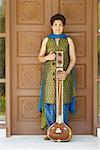 Portrait of a mature woman standing in front of a closed wooden door and holding a sitar