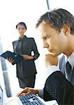 Businesswoman standing in backgroung, blurred, looking at businessman seated at computer, side view, close-up