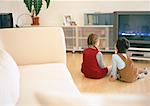 Lttle boy and girl sitting together on parquet floor looking at tv, rear view