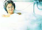 Woman smiling with eyes closed, listening to headphones in bubble bath