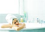 Woman in bathtub leaning on edge of tub, hair wrapped in towel