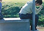 Teenage boy sitting on bench, his back turned to his friend holding his head, partial view