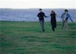 Three women, hand in hand, running across field of grass, sea on background