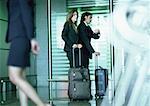Man and woman standing with suitcases, man looking at watch.