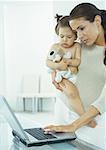 Woman holding little girl in one arm, typing on laptop with other hand