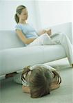 Little girl lying face down on floor, young woman sitting on sofa, reading