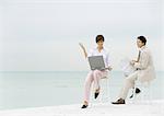 Two business people sitting on stools on beach, one using laptop, the other reading newspaper